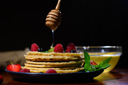Wooden stick in the form of a honeycomb with dripping honey on freshly baked pancakes for Shrovetide, served with ripe organic wild berries over a black background. Food still lifeの写真素材