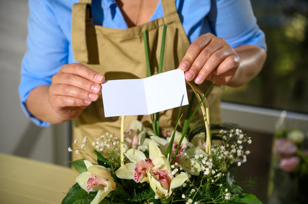 Close-up female florist holding white blank congratulatory card over a cute flower arrangement in a wicker basket. Gifts and floral arrangements in flower design studio. Copy advertising spaceの写真素材
