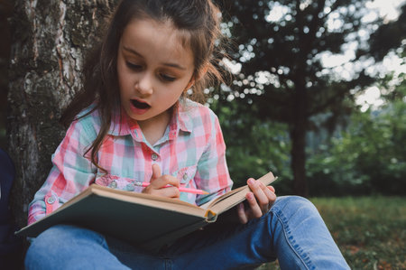Close-up portrait Caucasian beautiful little kid girl, first grader, primary school student, sitting under the tree and reading book, doing homework in the backyard after class. Education. Childhoodの写真素材