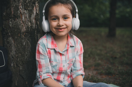 Close-up portrait of a Caucasian happy little child girl wearing wireless headphones, listening to music, relaxing after class, sitting under the tree on grass and cutely smiling looking at cameraの写真素材
