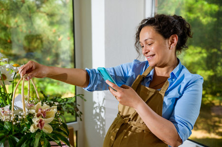 Smiling pregnant woman florist photographing a bouquet of orchid flowers in a wicker basket on her mobile phone, sharing it on social media, working in floral design studio. Arrangements and giftsの写真素材