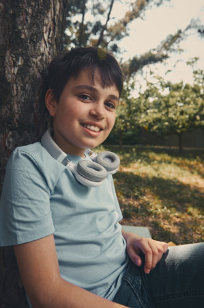 Close-up portrait of handsome adorable teenager, school boy with headphones, smiling looking at camera, sitting under a tree in the park. Toned portrait. People. Childhood. Pre adolescence. Lifestyleの写真素材