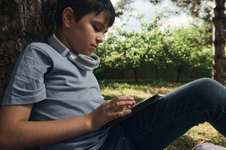 Close-up side toned portrait of a handsome serious smart teenage schoolboy 11-13 years old, primary school student, pupil with headphones, using digital tablet in park. Kids. Technology. Lifestyleの写真素材