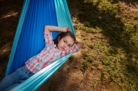 Top view Caucasian lovely little child girl relaxing outdoors, looking at camera, lying on a hammock on a sunny day, relaxing in summer camp or forest. Childhood. Recreation. Holidays. Vacations.の写真素材