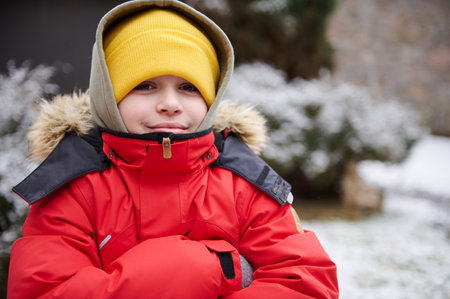 Close-up portrait of Caucasian handsome teenage boy 11-13 years old, in warm red down jacket and yellow wool hat, smiling looking at camera, standing in the snow covered backyard. Kids. Childhoodの写真素材