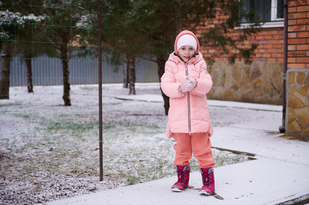 Full-length portrait of adorable little kid girl with rosy cheeks, wearing warm pink winter clothes, playing in a snowy backyard, smiling, looking at camera. Winter and christmas holidays conceptの写真素材