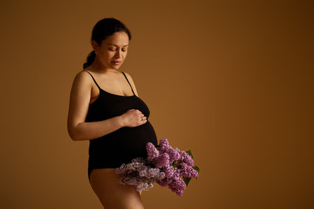 Attractive gorgeous young adult pregnant woman in black underwear, putting her hand on her big belly in late pregnancy, posing with a bunch of blooming lilacs, isolated over beige studio backgroundの写真素材