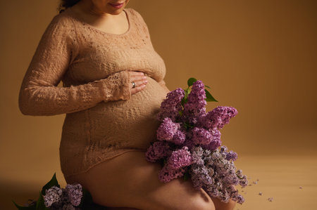 Midsection of a gravid woman in fine knitted elegant sweater putting her hand on her pregnant belly, sitting with a bunch of blooming fragrant lilac flowers, isolated on beige background. Copy spaceの写真素材