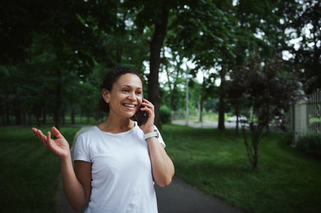 Beautiful multi-ethnic young woman in white t-shirt, smiling, talking on mobile phone while strolling the alley of a city park on a summer day. People. Communication. Wireless Technology. Lifestyleの写真素材