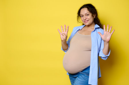 Cheerful multi-ethnic curly haired pregnant woman expecting a baby, showing the number nine with her...