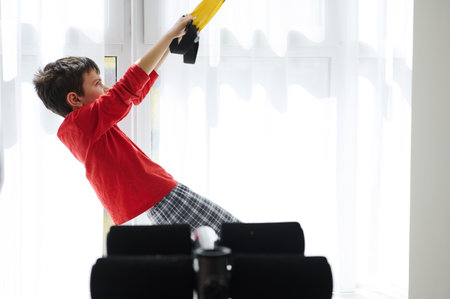 Side view of adorable Caucasian child, preteen boy exercising with suspension straps at home....