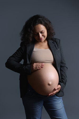 Stylish elegant multi-ethnic, curly haired pregnant woman smiling while gently caressing her big belly in pregnancy 36 week, feeling happy emotions from baby kicks, isolated on gray studio backgroundの写真素材