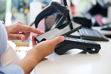 Close-up customer's hand holds modern smartphone over credit card reader, making contactless payments using NFC technology and mobile internet banking. People. Business. Convenience. Finance. Commerceの写真素材