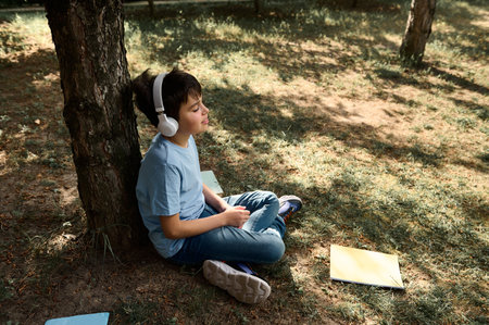 Happy Hispanic teenage boy in headphones, relaxing in the park after school, sitting under a tree...