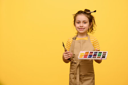 Confident creative little child, primary school girl holding watercolor palette, paint brush and palette knife, smiles cutely looking at camera, isolated on yellow studio background. Kids and hobbiesの写真素材