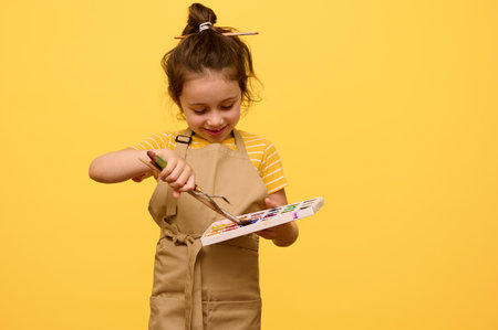 Smiling little child girl artist, schoolgirl dressed as an artist in beige apron, learning painting art, dipping paintbrush into a palette with watercolor paints, isolated yellow backdrop. Copy spaceの写真素材