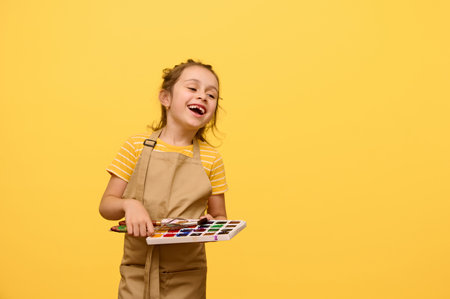 Beautiful Caucasian little child girl laughing, expressing happy positive emotions during art class, holding a palette with watercolor paints and paintbrush, isolated over yellow studio backgroundの写真素材