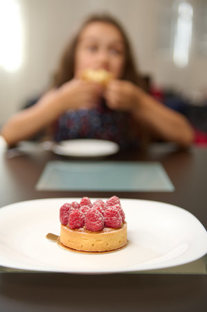 Close-up of a tasty sweet French dessert - a tartlet with raspberries on white plate, over blurred background of a little child taking her breakfast or snacking. Food still life. Culinary. Patisserieの写真素材