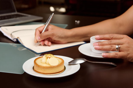 Close-up of woman's hand using a stylish pen, writing on notepad, planning project, enjoying her coffee break with delicious tasty French dessert, a lemon tartlet. Business conceptの写真素材