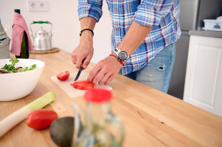 Close-up of the hands of a male chef, a cook cutting fresh ripe organic juicy tomatoes while preparing salad in the home kitchen. Healthy eating and dieting concept. People. Lifestyle. Food and drinkの写真素材