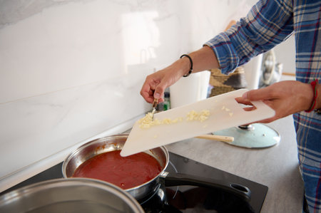 Close-up of chef's hands putting chopped garlic from a cutting board into a saucepan with boiling tomato sauce. People and good conceptの写真素材