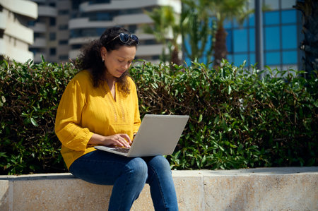 Confident multi-ethnic young adult female student working online on a laptop, sitting on a stone bench against urban buildings background. People. Internet business. Education and career conceptの写真素材
