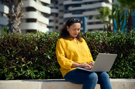Confident purposeful multi-ethnic young adult businesswoman telecommuting, using laptop for remote work outdoors. People. Education. Career. Online business concept. Female entrepreneur using computerの写真素材