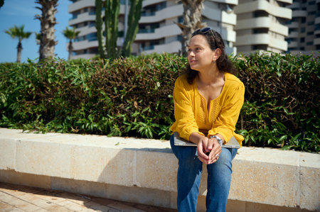 Portrait of a confident pensive authentic pretty woman holding laptop on her knees, sitting on a stone bench outdoor and thoughtfully looking aside, thinking on new startup, planning business projectの写真素材