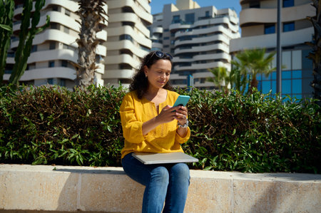Beautiful multi-ethnic freelance woman with laptop, using smart mobile phone, booking, ordering food, resting outdoor against high rise modern buildings background. People. Technology. Businessの写真素材