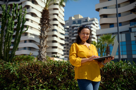 Authentic business woman, real estate agent holding laptop, smiling looking confidently at camera, standing against high rise buildings background, dressed in casual clothes. People and businessの写真素材