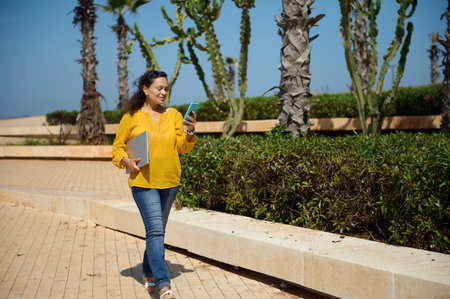 Full length portrait of a pretty woman in casual clothes checking her smartphone, carrying a laptop while walking on the street against palm trees background on a warm sunny day. People. Career. Jobの写真素材