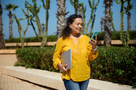 Middle Eastern confident beautiful female entrepreneur, business woman holding a laptop, checking mobile application on her modern smartphone, strolling the coastline street on a warm sunny summer dayの写真素材