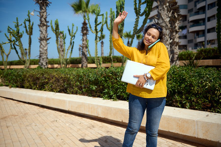 Smiling multitasking female entrepreneur holding laptop, talks on mobile phone and greeting her colleagues, standing in corporate meeting place, against high rise buildings background. People. Careerの写真素材