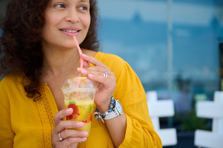 Close-up portrait of a young multi-ethnic beautiful woman drinking fresh juice outdoor, pensively looking aside a copy advertising space on glass windows. People. Lifestyle. Food and drink consumerismの写真素材