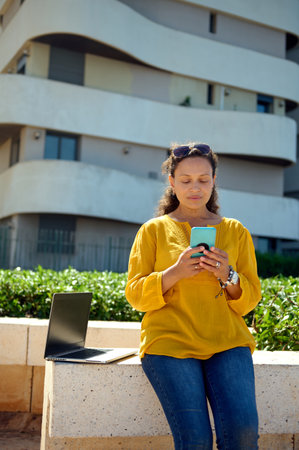 Beautiful woman using smart mobile phone, checking new application, standing near her laptop with black screen outdoor. Portrait of sales manager, freelancer online working, dressed in casual clothesの写真素材