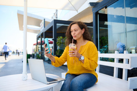 Beautiful woman, blogger drinking orange juice and taking selfie, sharing her lifestyle on social media content. Freelancer working online on laptop outdoors. People. Business. Leisure. Bloggingの写真素材