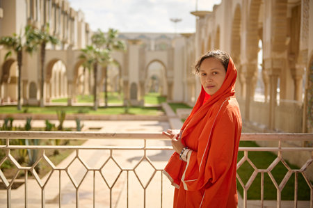 Charming ethnic woman in religious outfit and head covered in orange scarf, looking confidently at the camera, standing on the balcony against the background of marble arches and garden in the mosqueの写真素材