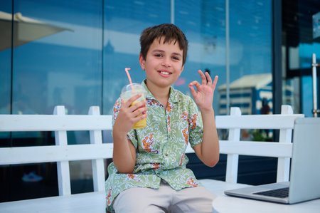 Handsome schoolboy in summer shirt, holds a glass of freshy squeezed orange juice, shows OK hand...