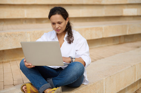Multi ethnic young adult businesswoman, entrepreneur freelance worker working on laptop while sitting on steps. People. Business. Career. Internet technology. E-learning and online education conceptの写真素材