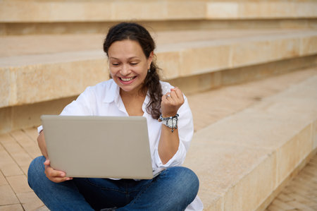 Happy Latina business woman clenching fists while online working on laptop, sitting outdoors on steps. Goals achievement. Success. people. Career and recruitment. Online communication and remote workの写真素材