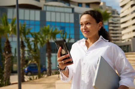 Portrait of young smiling dark-haired woman in casual white shirt walking city streets and using smartphone to interact with her business partners. People. Communication. Modern technologies conceptの写真素材