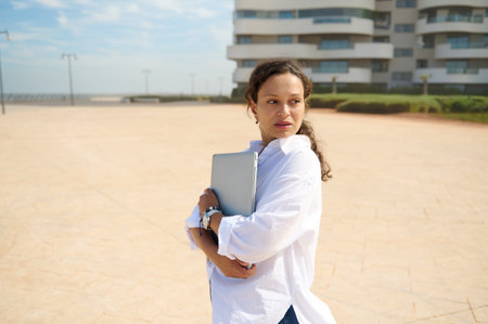 Pensive young adult pretty woman holding laptop, thoughtfully looking aside, standing outdoors against modern high rise buildings background. Real estate investment. Online business. People and careerの写真素材