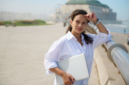 Portrait of beautiful curly haired woman, freelancer with laptop standing against mosque background, holding a laptop and thoughtfully looking into the distance. People, career and business conceptの写真素材