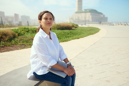 Confident business woman, freelance entrepreneur sitting on bench outdoors against a Hassan II mosque background, thinking on new startup project, dreamily looking into the distance. People. Lifestyleの写真素材