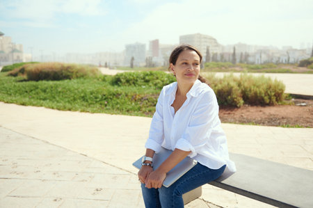 Attractive successful brunette, business woman, manager, office worker, smiles looking aside, sitting outdoors against cityscape backdrop. People Lifestyle Remote work. Career and recruitment conceptの写真素材