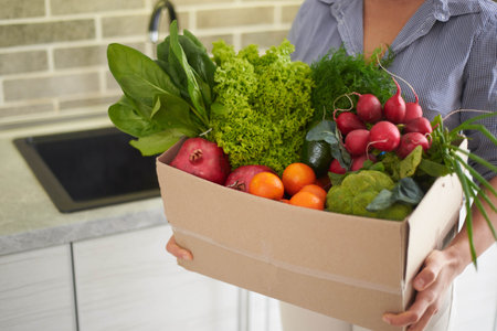 Close-up of a cardboard box full of fresh organic vegetables, fruits, herbs and greens in female hands, standing in the kitchen at home. Housewife carrying a recyclable container of raw vegan foodの写真素材