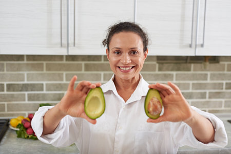 Multi ethnic smiling woman looking at camera and showing two halves of fresh ripe organic avocado...