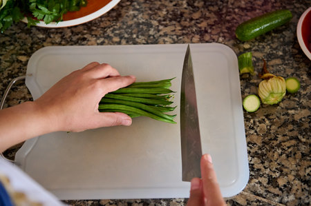 Top view of a housewife, woman chef holding a kitchen knife, chopping fresh green beans on a cutting board, standing by marble kitchen counter in the home kitchen. Raw food. Vegetarianism. Close-upの写真素材