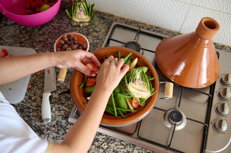 View from above of Muslim housewife cooking delicious vegetarian meal in tagine clay pot, standing by kitchen counter at home, according to traditional Moroccan recipe. Culinary and culture in Moroccoの写真素材