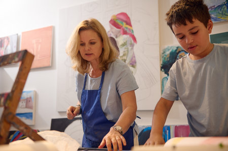 Adorable Caucasian teenage school boy learning painting and art, standing with a female paint teacher artist by a desk, working together in creative visual art workshopの写真素材